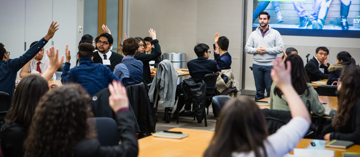 A man stands in front of a classroom full of students raising their hands.
