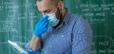 A man with gloves and a mask on in front of blackboard touching his mask and holding a notepad