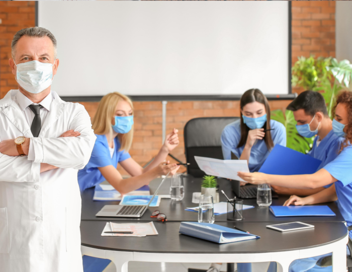 A doctor stands with his arms crosed and a mask on. In the back four nurses, three women and one man, with masks on discussing something