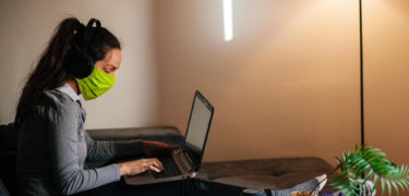 A woman wearing headphones and a mask sitting on the couch while typing on her computer.