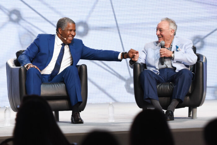 Photo of Robert F. Smith and Sanford I. Weill, who are both sitting on stage during a speaking engagement. Robert F. Smith is wearing a blue suit and a white collared shirt. Sanford I. Weill is wearing a blue suit and a gray shirt and holding a microphone. The two men are in the process of greeting each other.