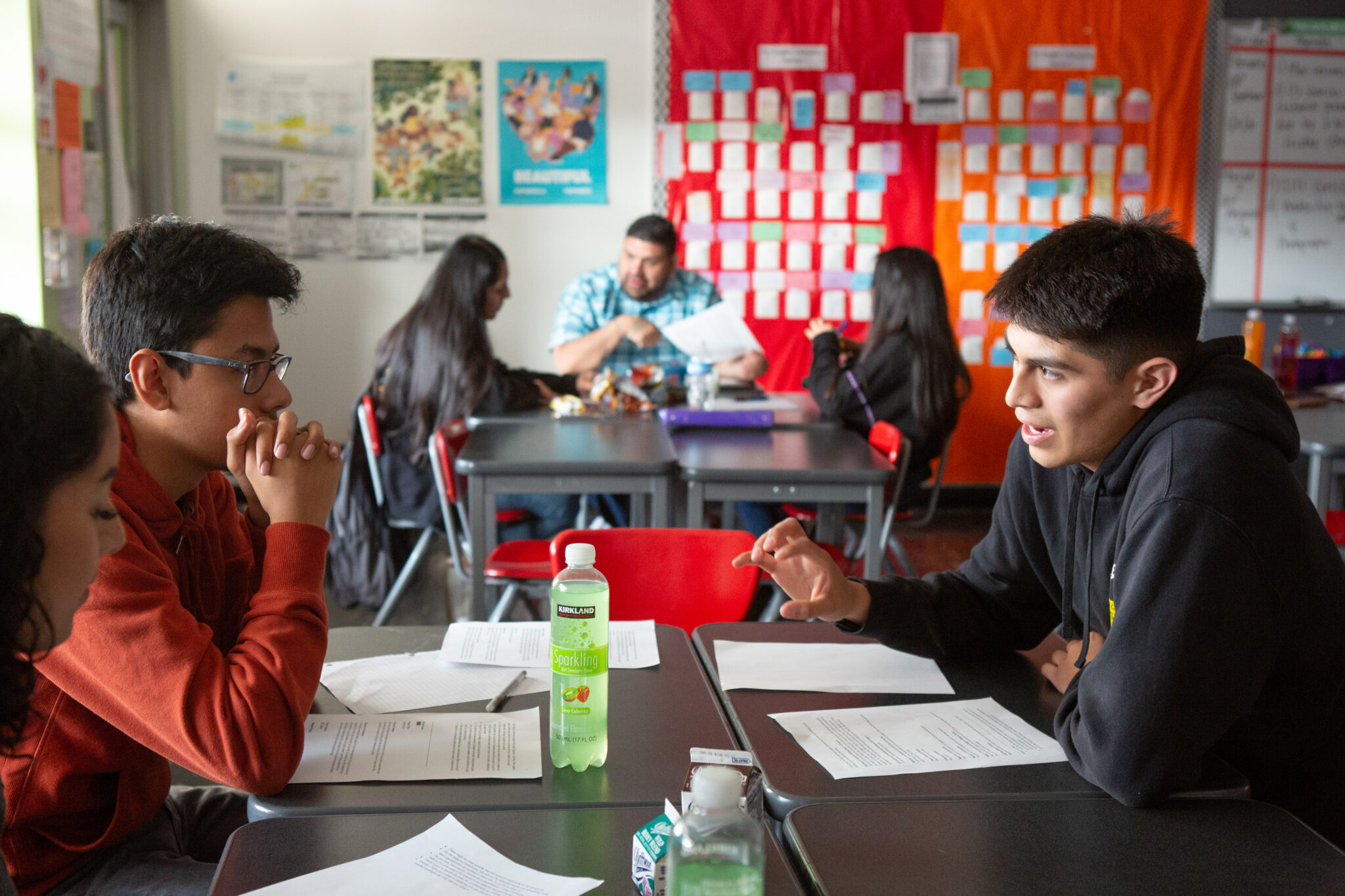 Students engage in group discussion in a classroom. The background features colorful bulletin boards.